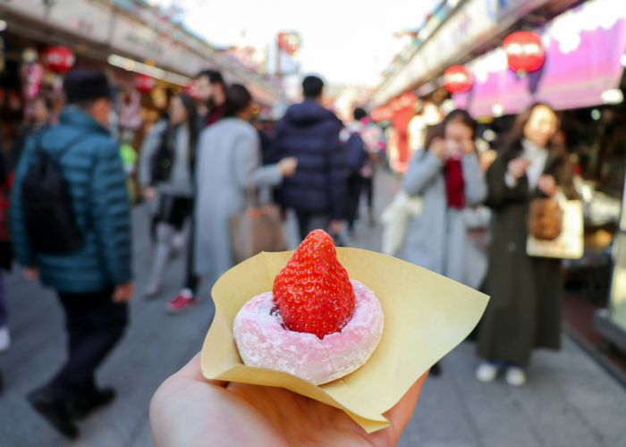 A strawberry daifuku held in the foreground, with a busy market street in the background.